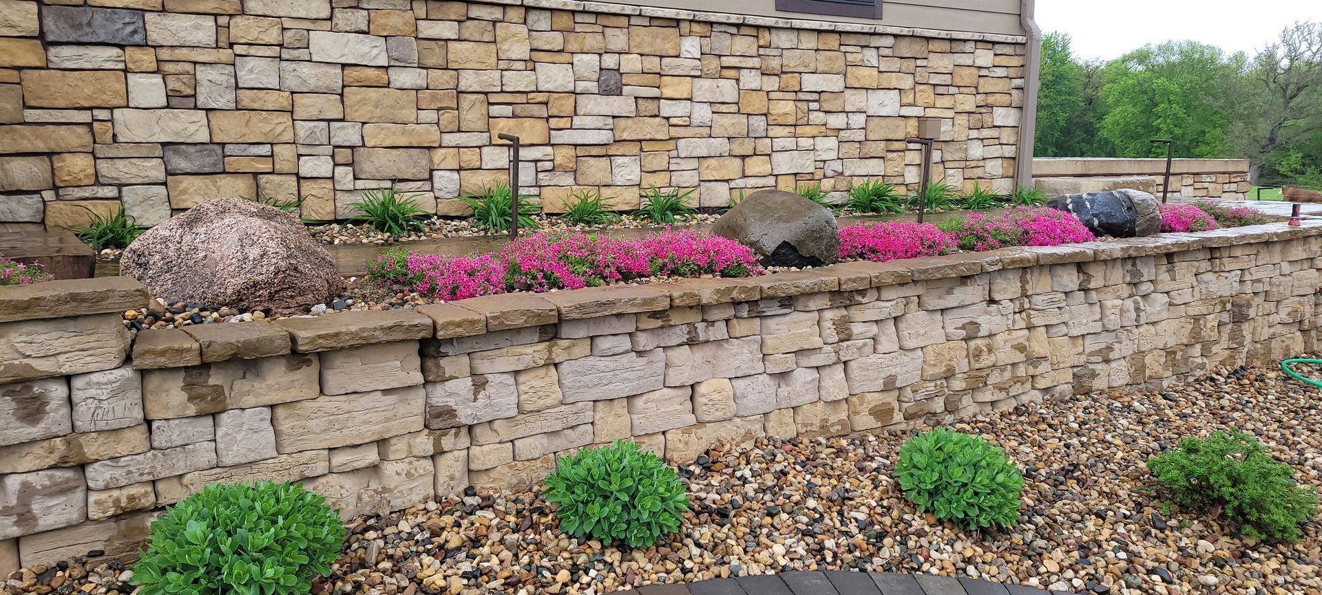 A low, stacked stone retaining wall with a landscaped bed featuring purple flowers, dark rocks, and green shrubs.