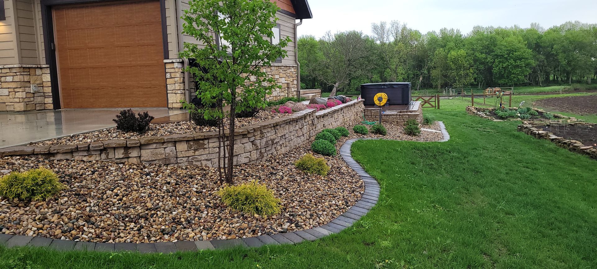 A landscaped gravel garden bed with shrubs and a small tree, bordered by curved stone pavers next to a suburban home.
