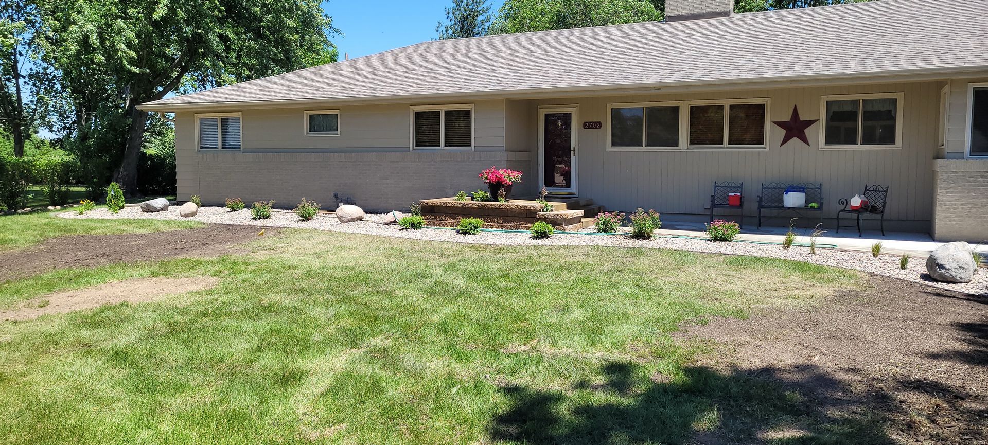 A beige, single-story ranch house with a gray shingled roof, a front porch, and a landscaped yard.