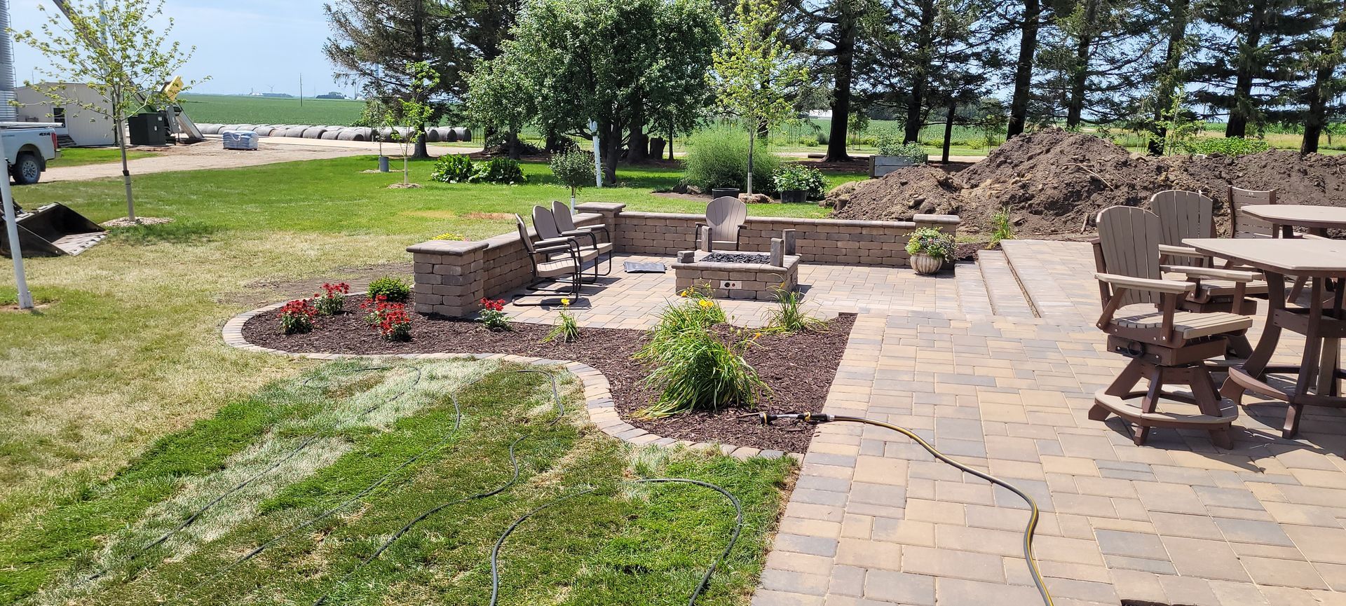 A backyard patio with a stone fire pit, seating, and a garden bed against a backdrop of trees and a grassy lawn.
