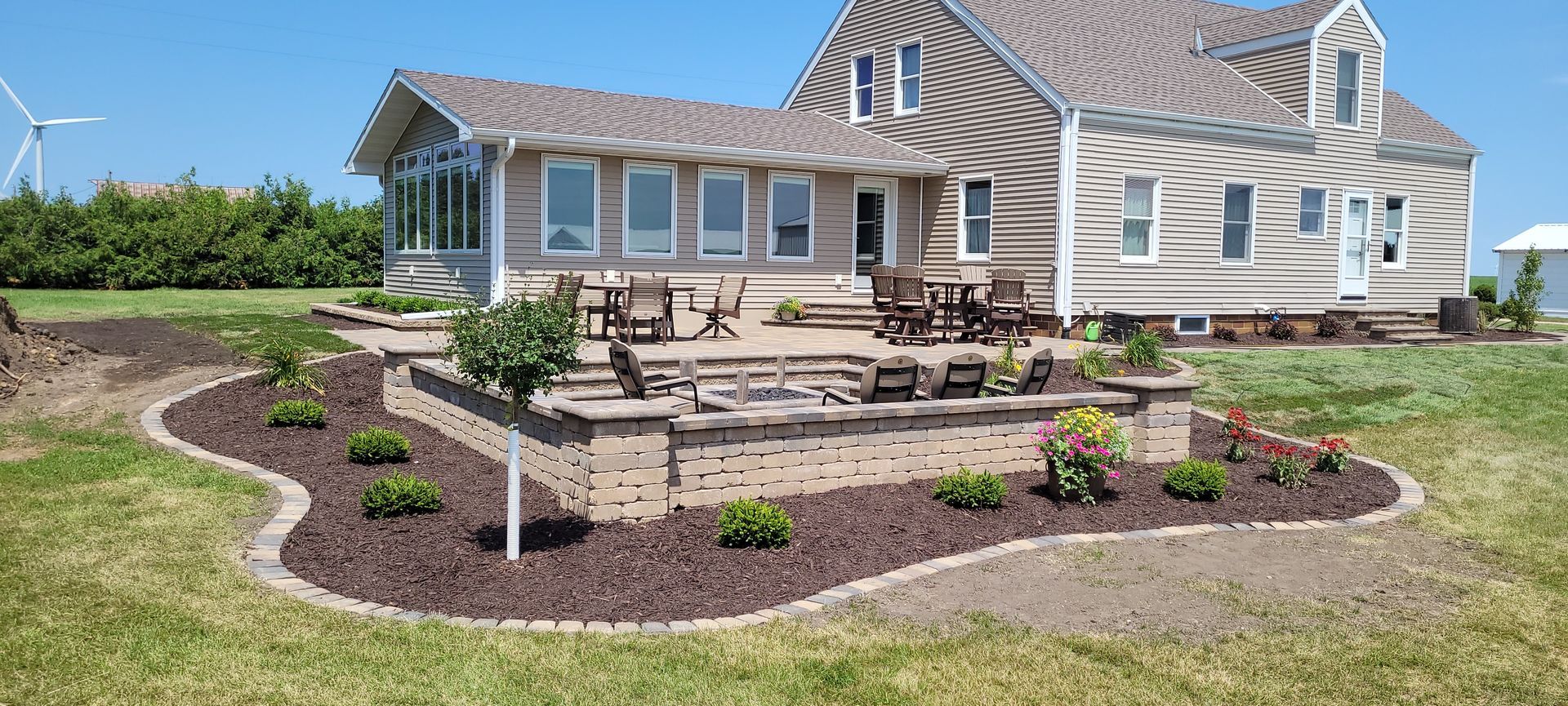 A paved patio with a fire pit, outdoor furniture, and brick retaining walls sits beside a tan house under a blue sky.