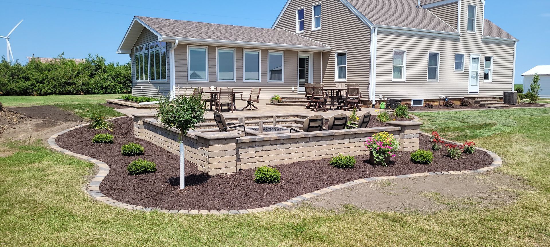 A beige house with a stone patio, outdoor seating, and a landscaped mulch garden with small shrubs in a grassy yard.