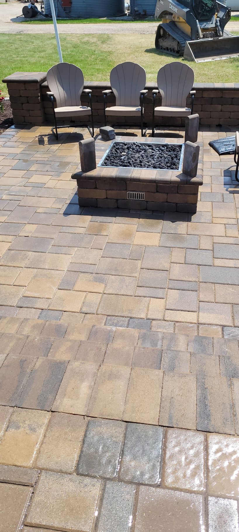 A paver patio with a stone fire pit and three tan Adirondack chairs arranged against a stone retaining wall.