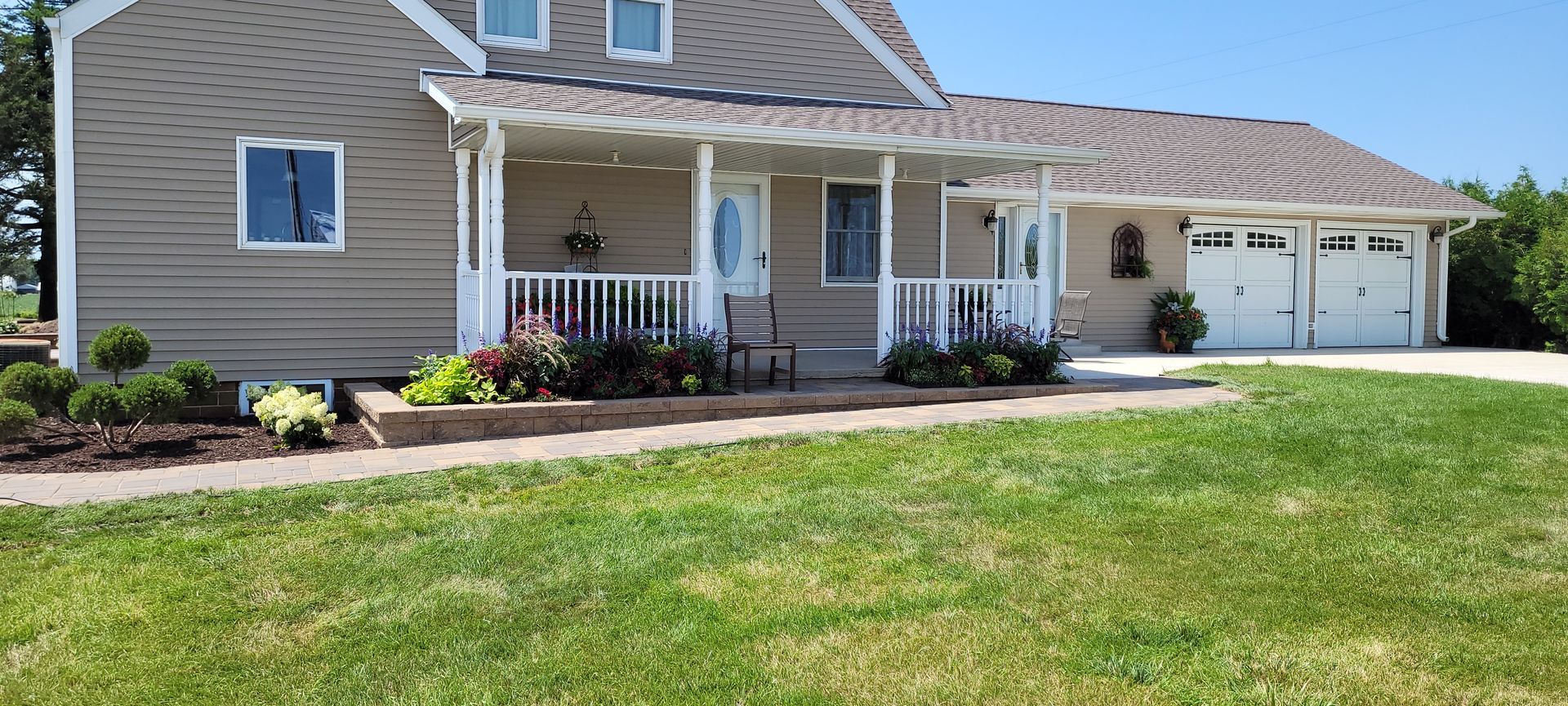 Tan house with a covered front porch, landscaping, and a two-car garage on a sunny day.