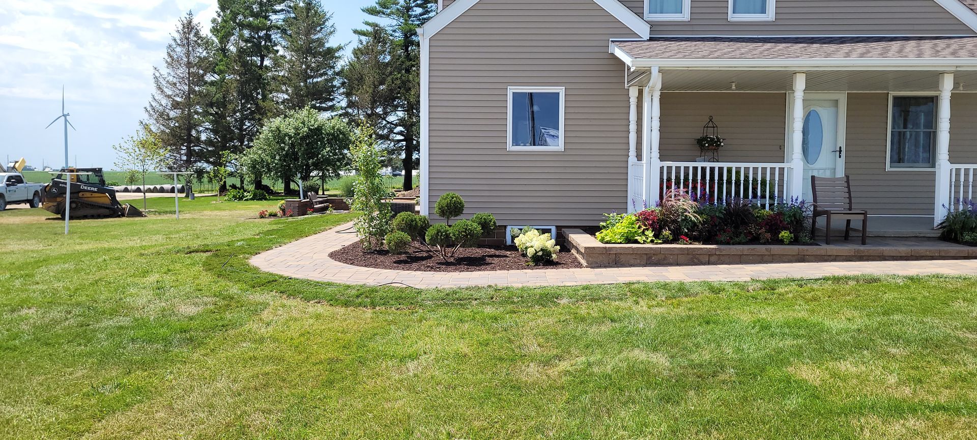 A beige house with a wrap-around porch and foundation garden, set beside a grassy lawn and a distant field.