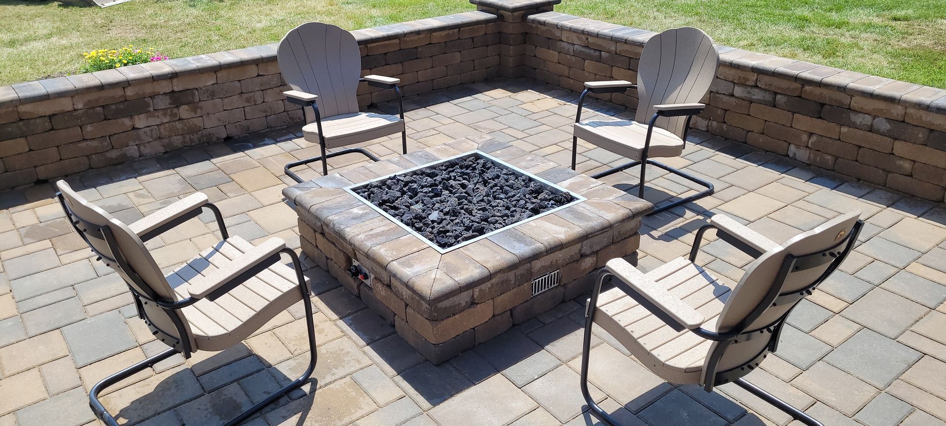 A square stone fire pit surrounded by four beige patio chairs on a paved patio in a backyard.