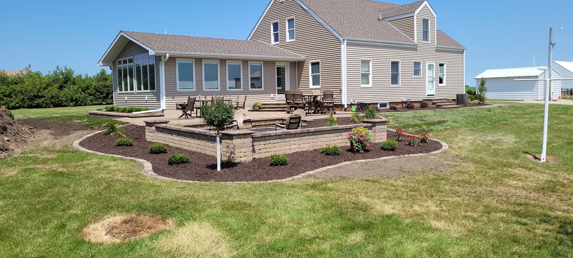 A landscape view of a tan, multi-story home with a wrap-around porch and a raised stone patio surrounded by fresh mulch.