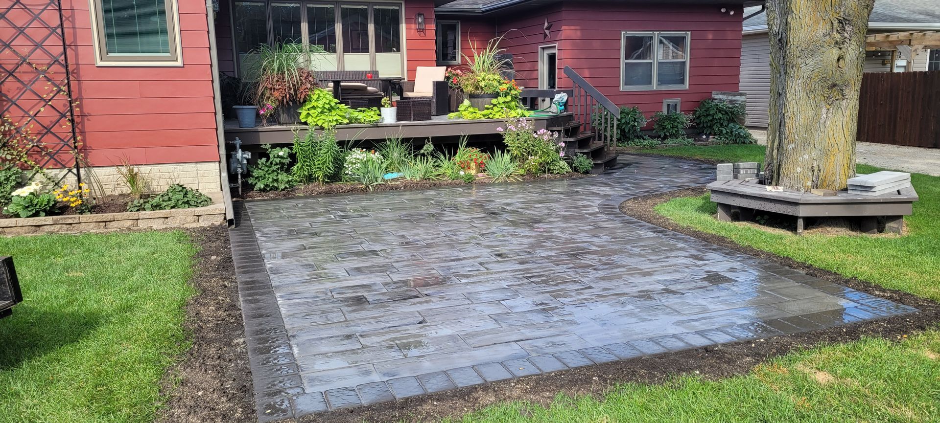 A newly installed gray stone patio sits in a backyard next to a red house, wooden deck, and a tree with a bench surround.