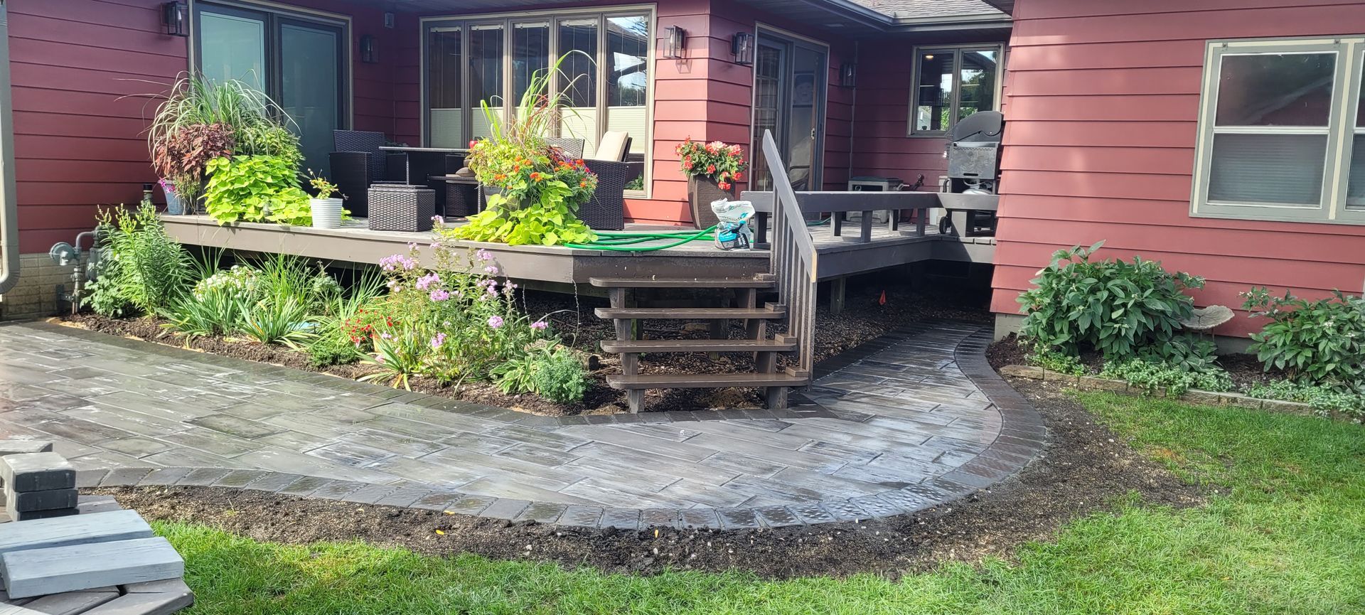 A red house with a wooden deck featuring potted plants and stairs leading to a curved stone patio and lawn area.