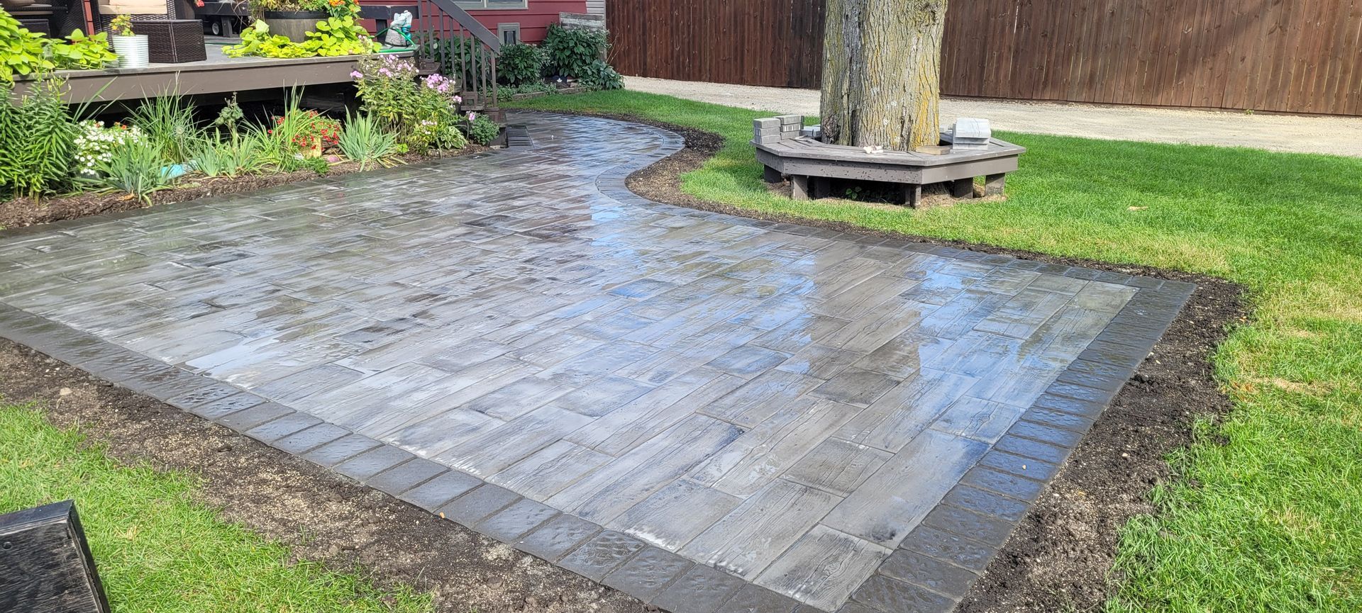 A freshly laid grey stone patio with a dark border, situated in a grassy backyard next to a tree with a bench.