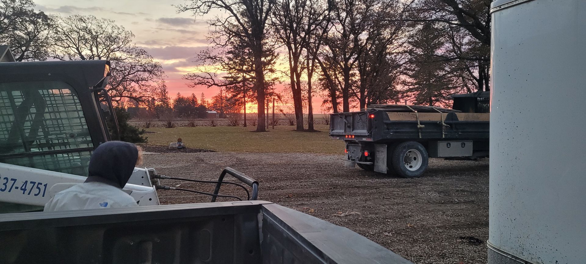 A person in a hooded jacket sits near a tractor as a truck stands on a dirt field under a sunset sky.