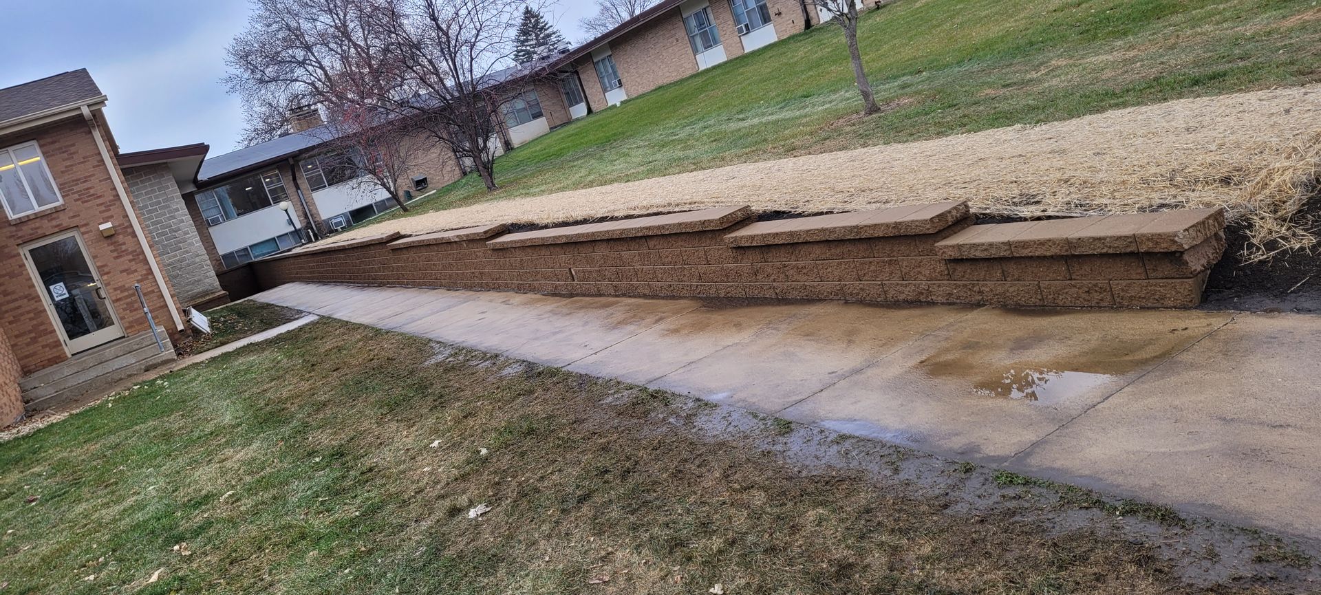 A low, stacked stone retaining wall runs alongside a paved walkway next to a brick building and a sloped grassy hill.