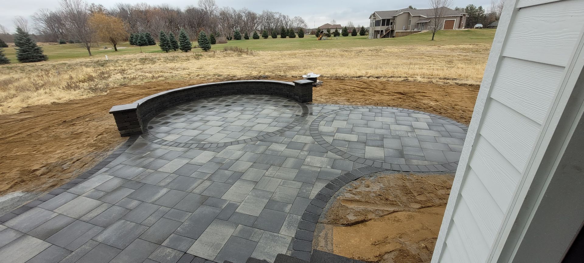 A newly installed curved gray stone patio with a retaining wall, situated in a large backyard near a house.