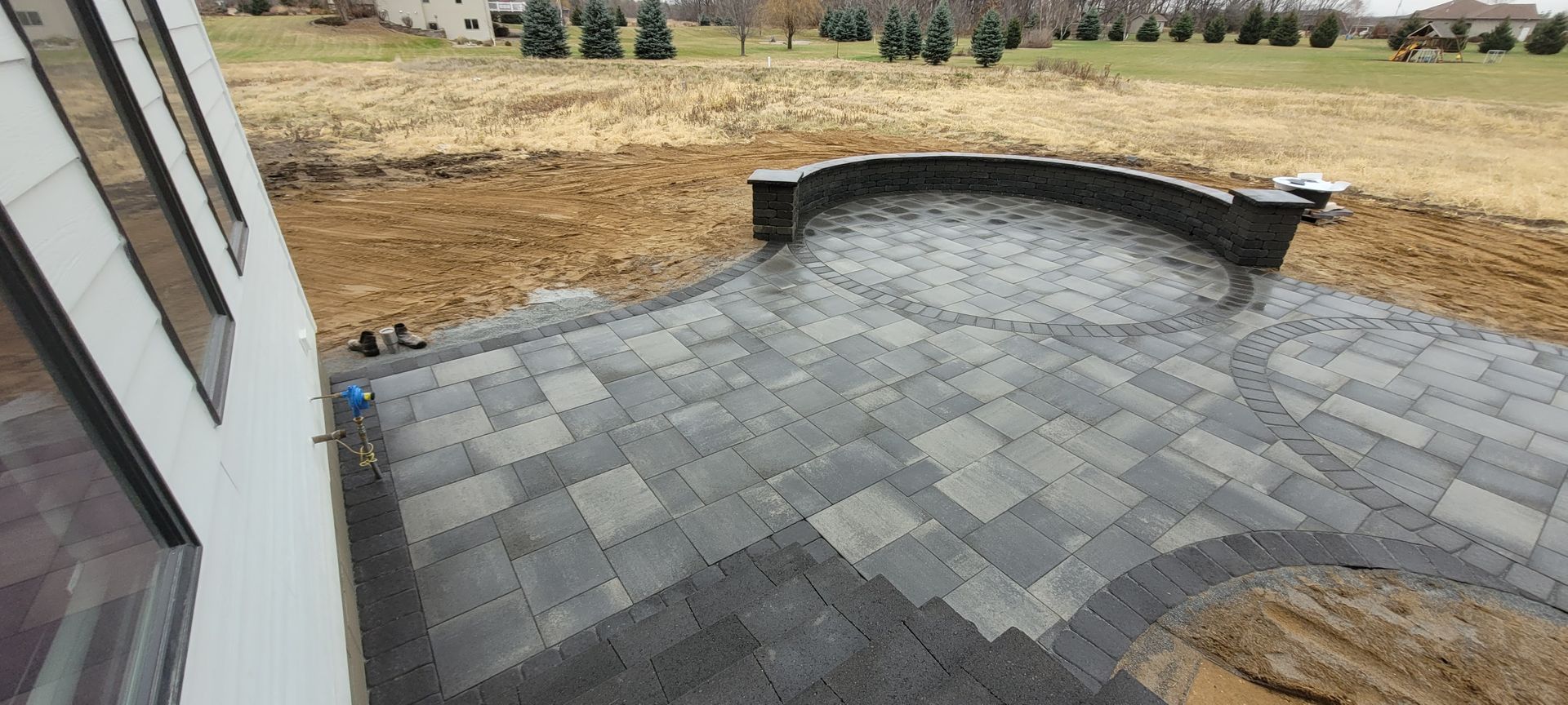 A newly installed gray paver patio with a curved seating wall, bordered by exposed soil against a house exterior.