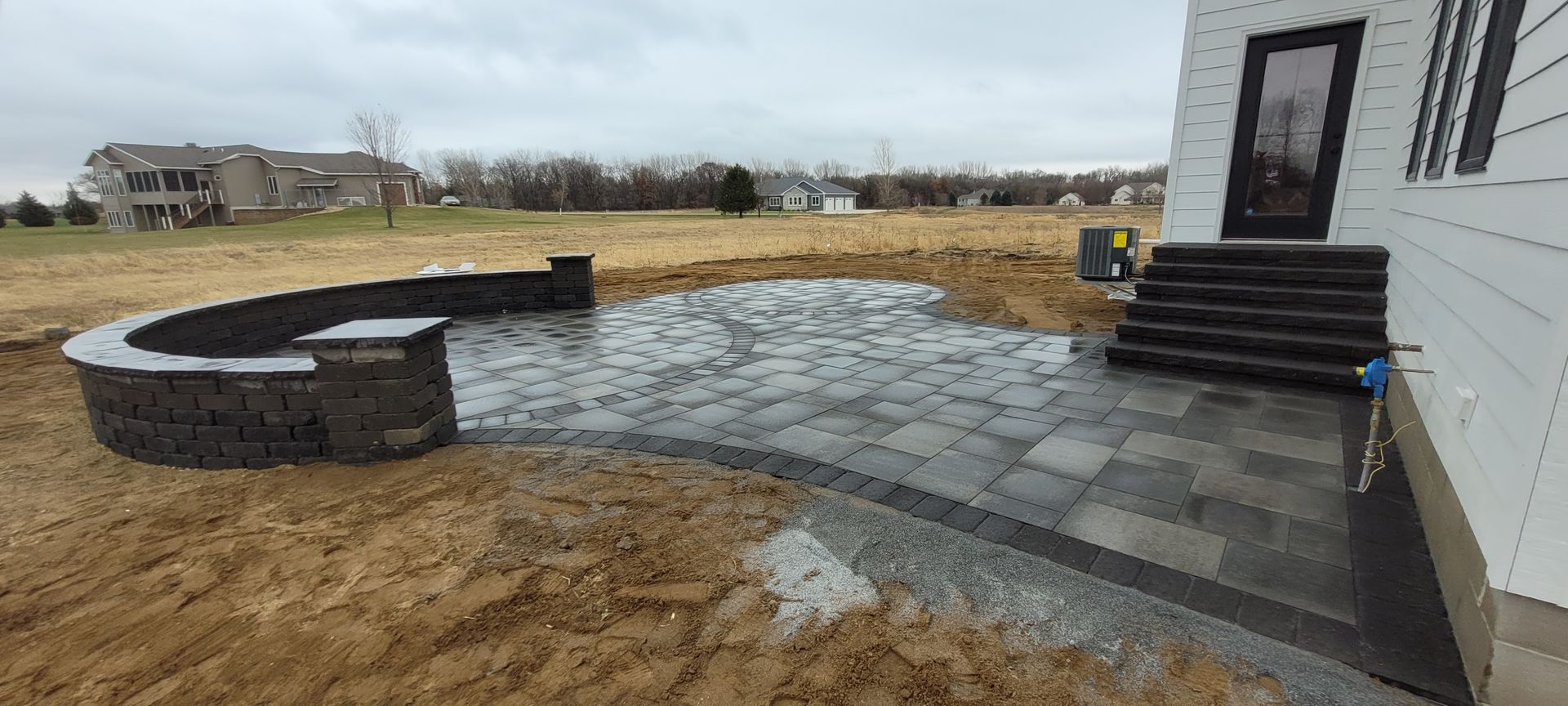 A newly installed paver patio with a curved stone retaining wall and dark steps leading to a white house.