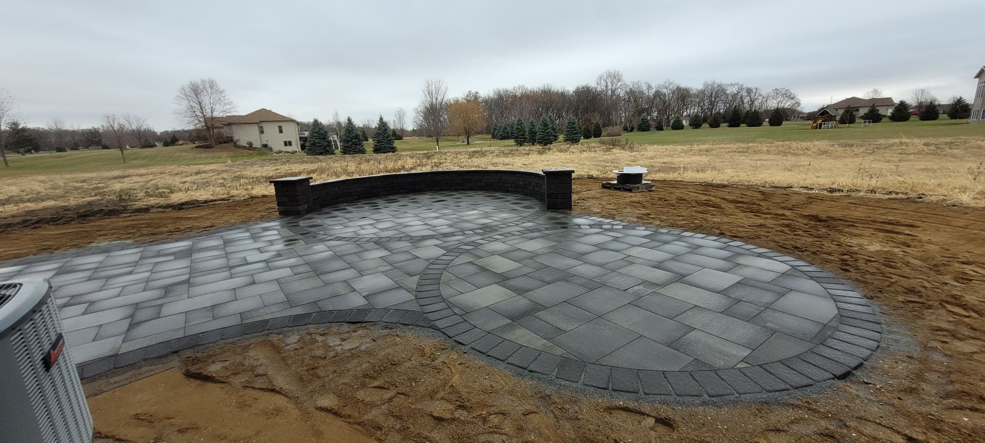 A newly installed grey stone patio with a curved retaining wall sits in a backyard on an overcast day.