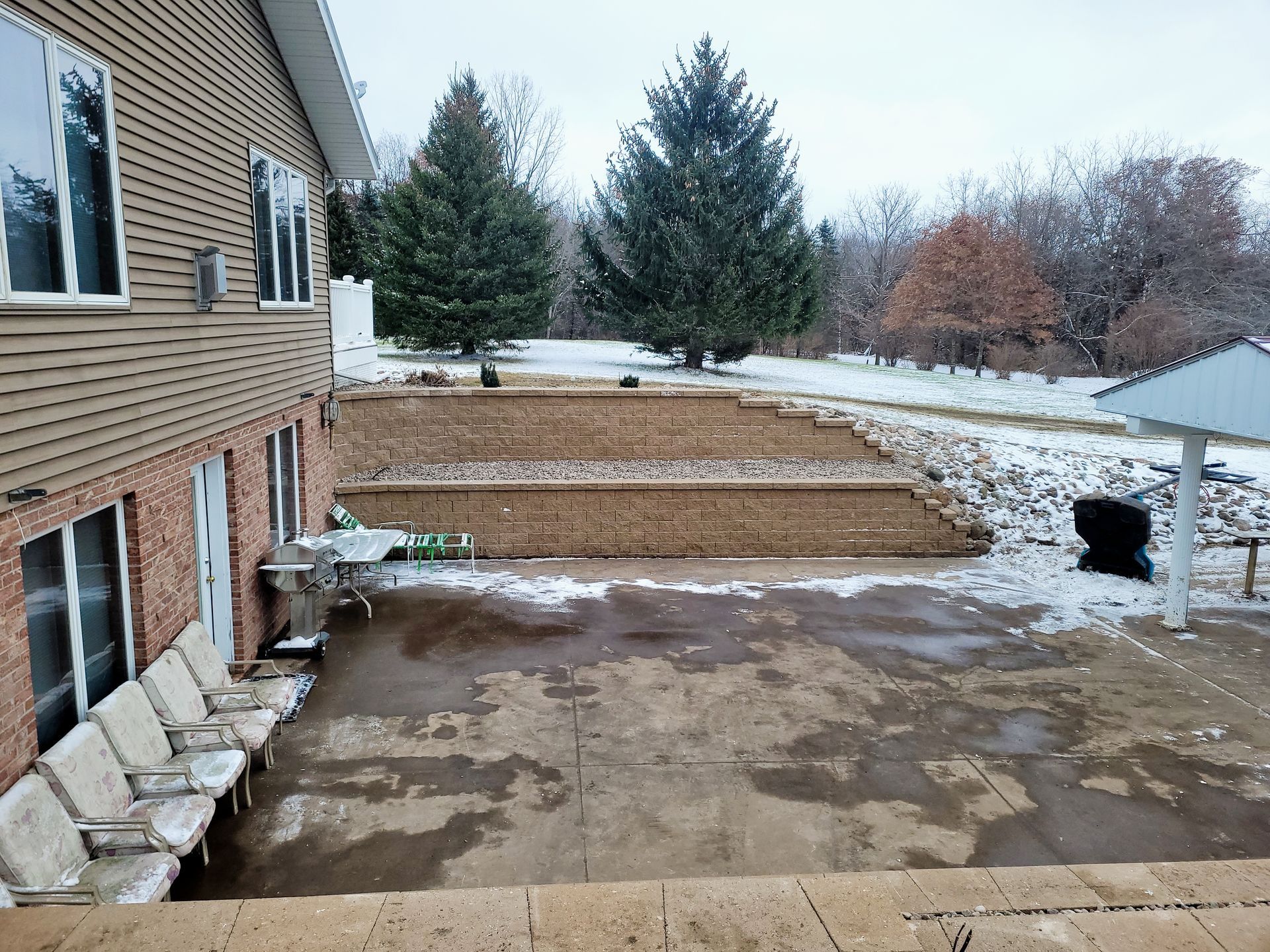 A side view of a house with a concrete patio, lined chairs, and a tiered stone retaining wall in a snow-dusted backyard.