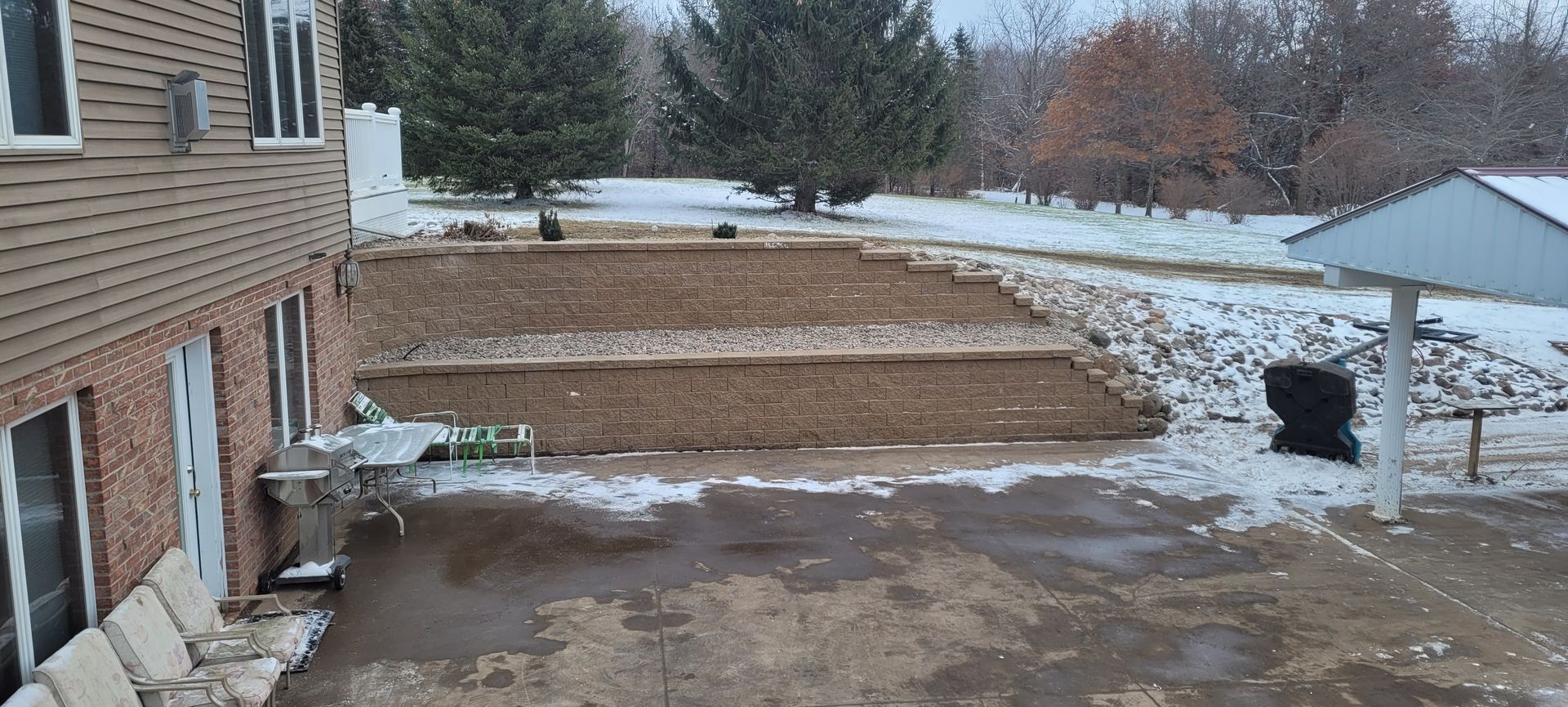 A tiered landscape of tan stone retaining walls in a snow-covered backyard next to a brick house.