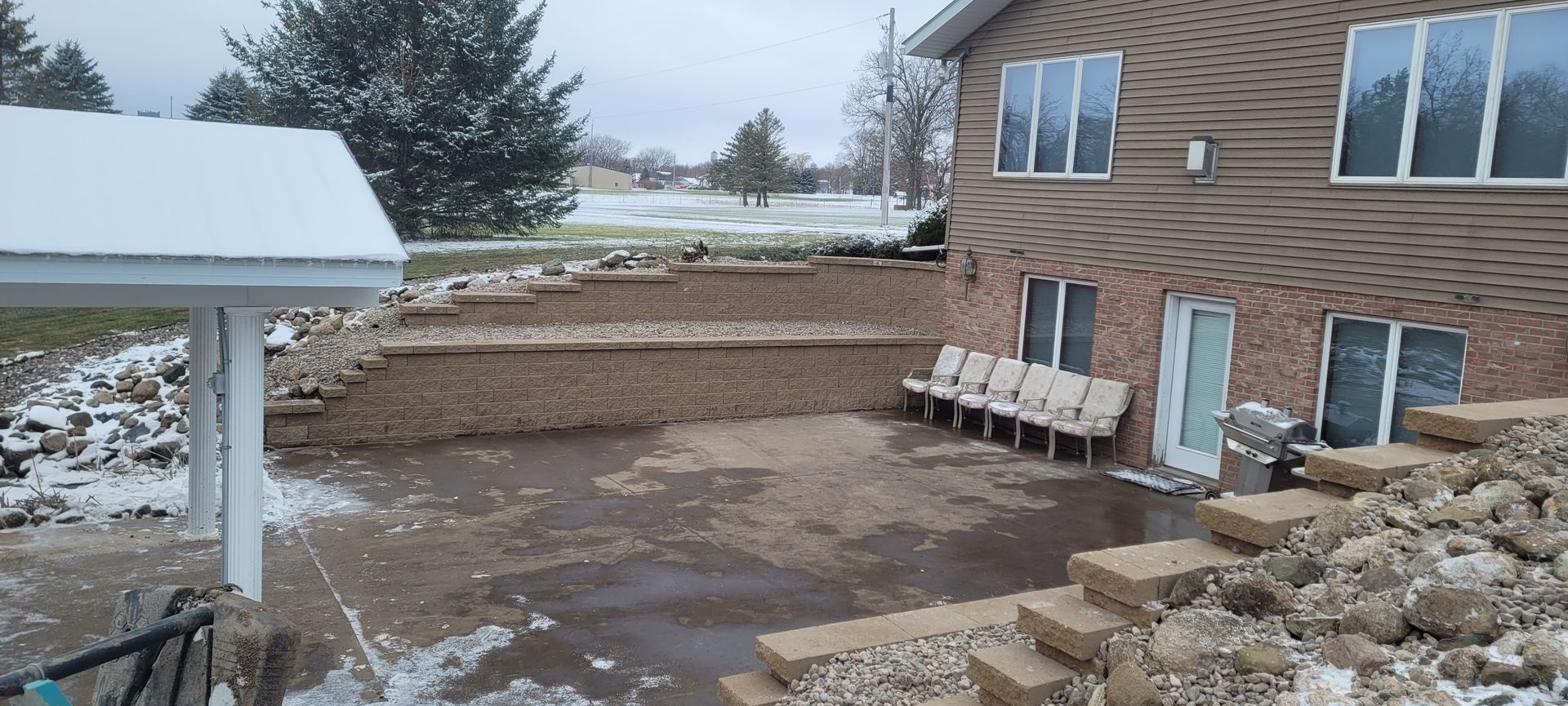A multi-level backyard patio with stone retaining walls, a covered porch, and light snow on the ground near a brick house.