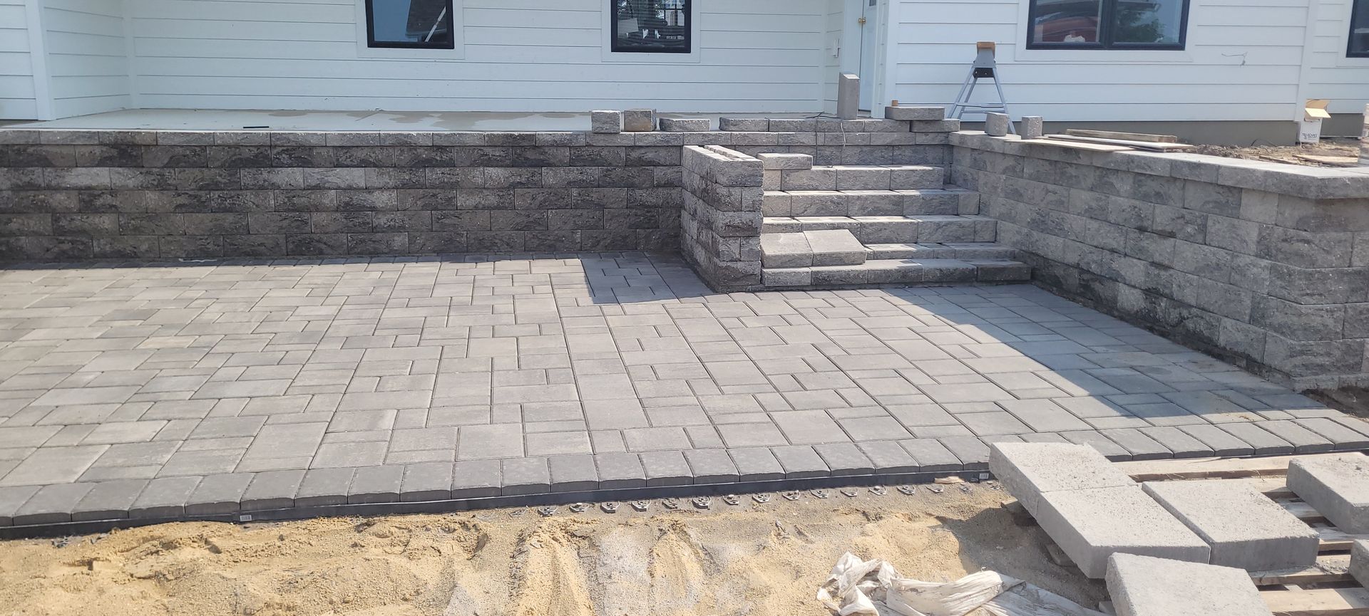 A newly constructed gray paver patio and retaining wall with stone steps leading to the back of a white house.