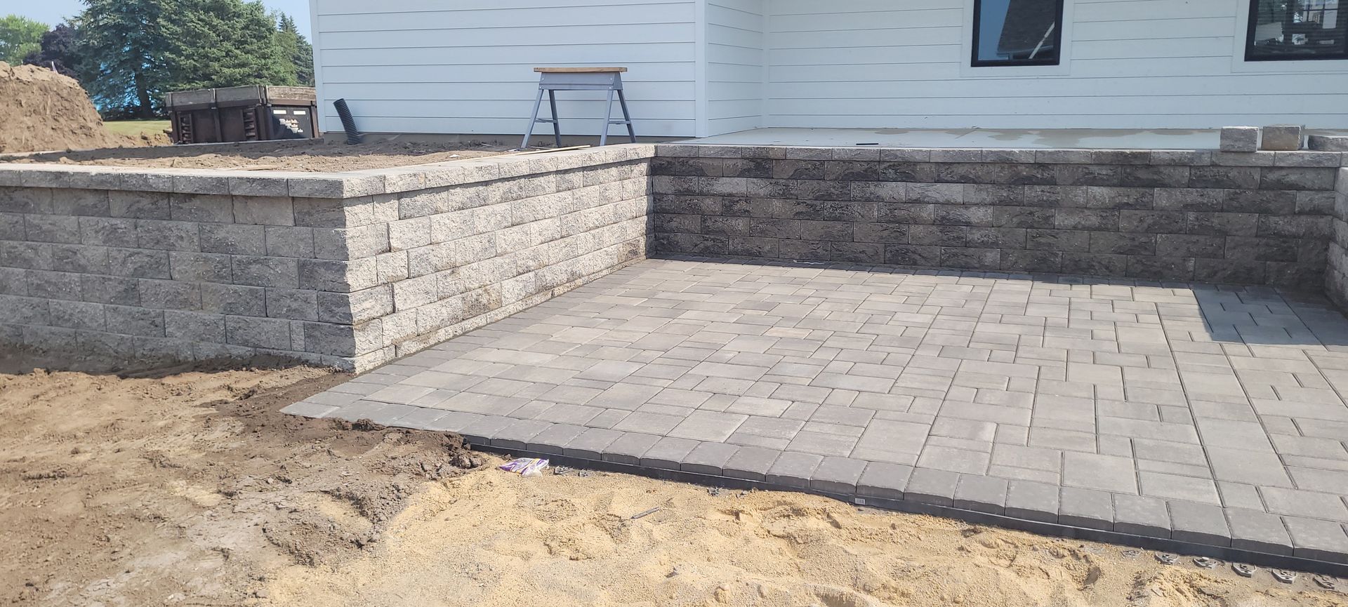 A gray paver patio sits adjacent to a white house with a multi-tiered retaining wall made of textured stone blocks.