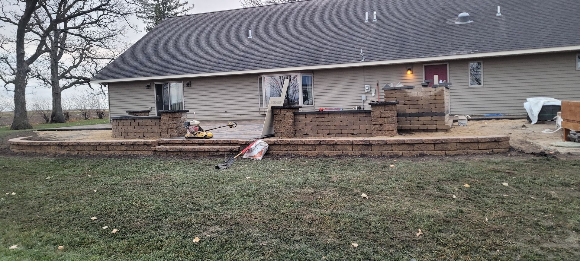 A backyard under construction featuring a stone retaining wall and patio area attached to a tan siding house.