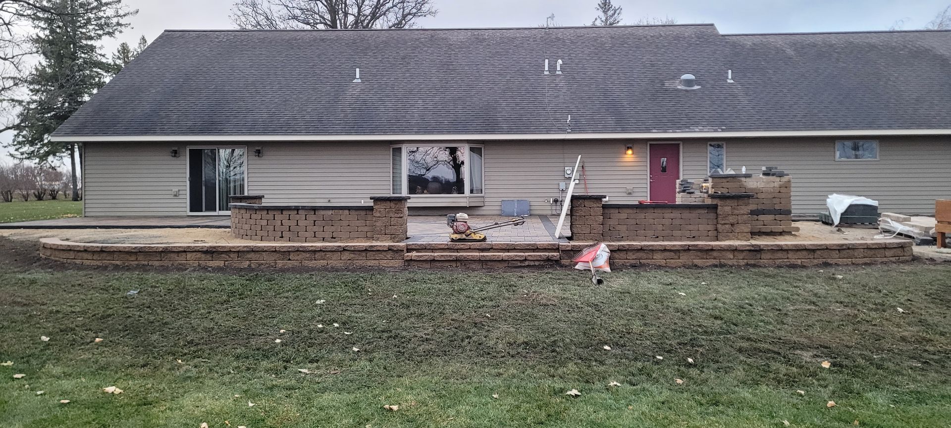 A ranch-style house with a tan exterior and a brick patio under construction in the backyard.