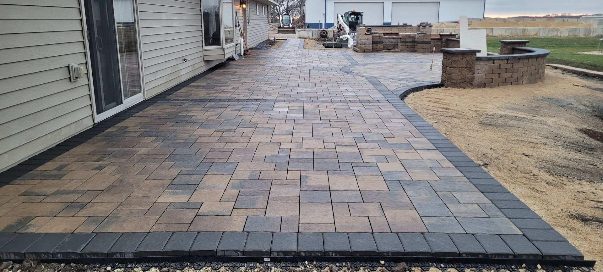 A newly installed outdoor paver patio featuring tan and grey stones with a dark grey border, next to a house siding wall.