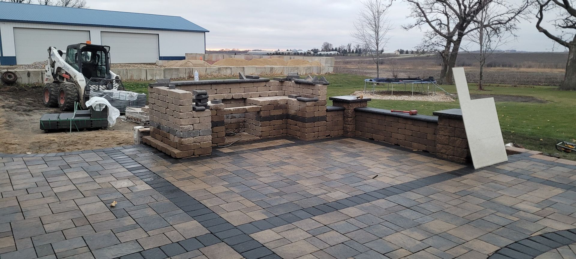 A partially constructed outdoor kitchen with brick walls and stone patio pavers on a rural property with a skid steer.