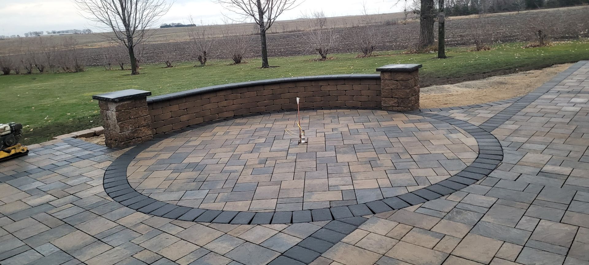 A circular patio made of patterned tan and grey pavers with a curved stone retaining wall and two matching pillars.