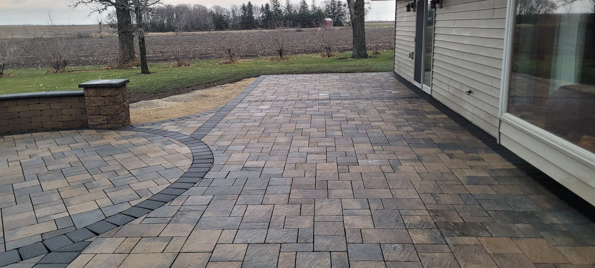 A patio made of grey and brown rectangular pavers, featuring a circular design, extending from a house toward a field.