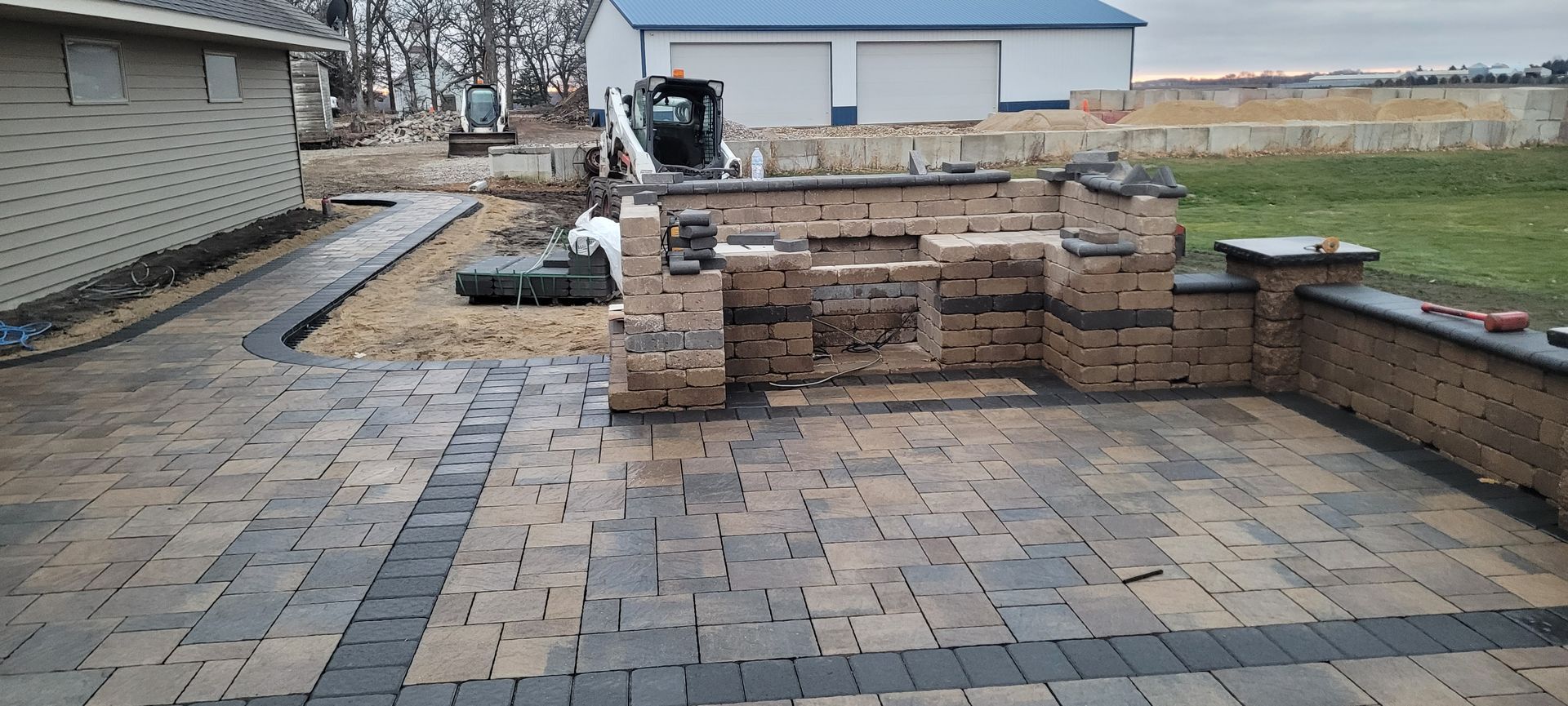 A partially finished backyard patio featuring tan and gray pavers, a brick outdoor kitchen, and a sidewalk along a house.