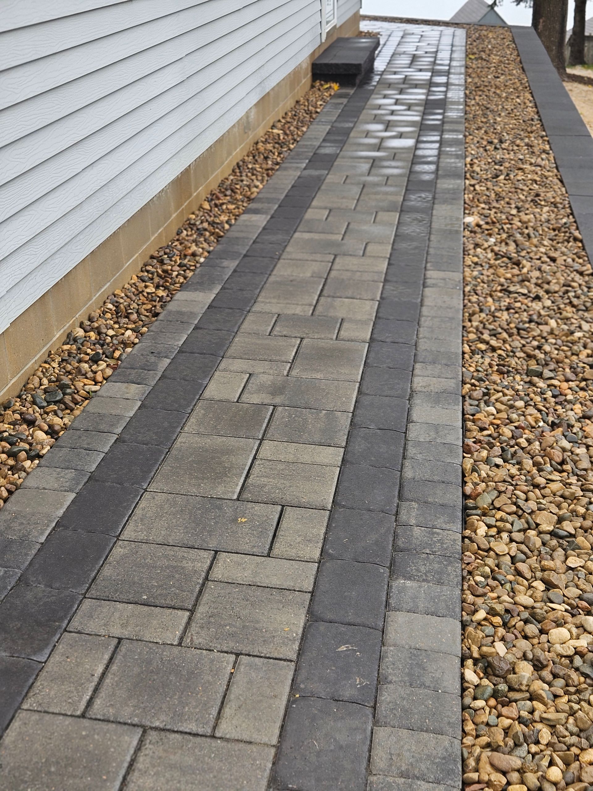 A paved walkway with gray, multi-sized stone pavers, bordered by dark charcoal bricks and loose tan gravel on both sides.