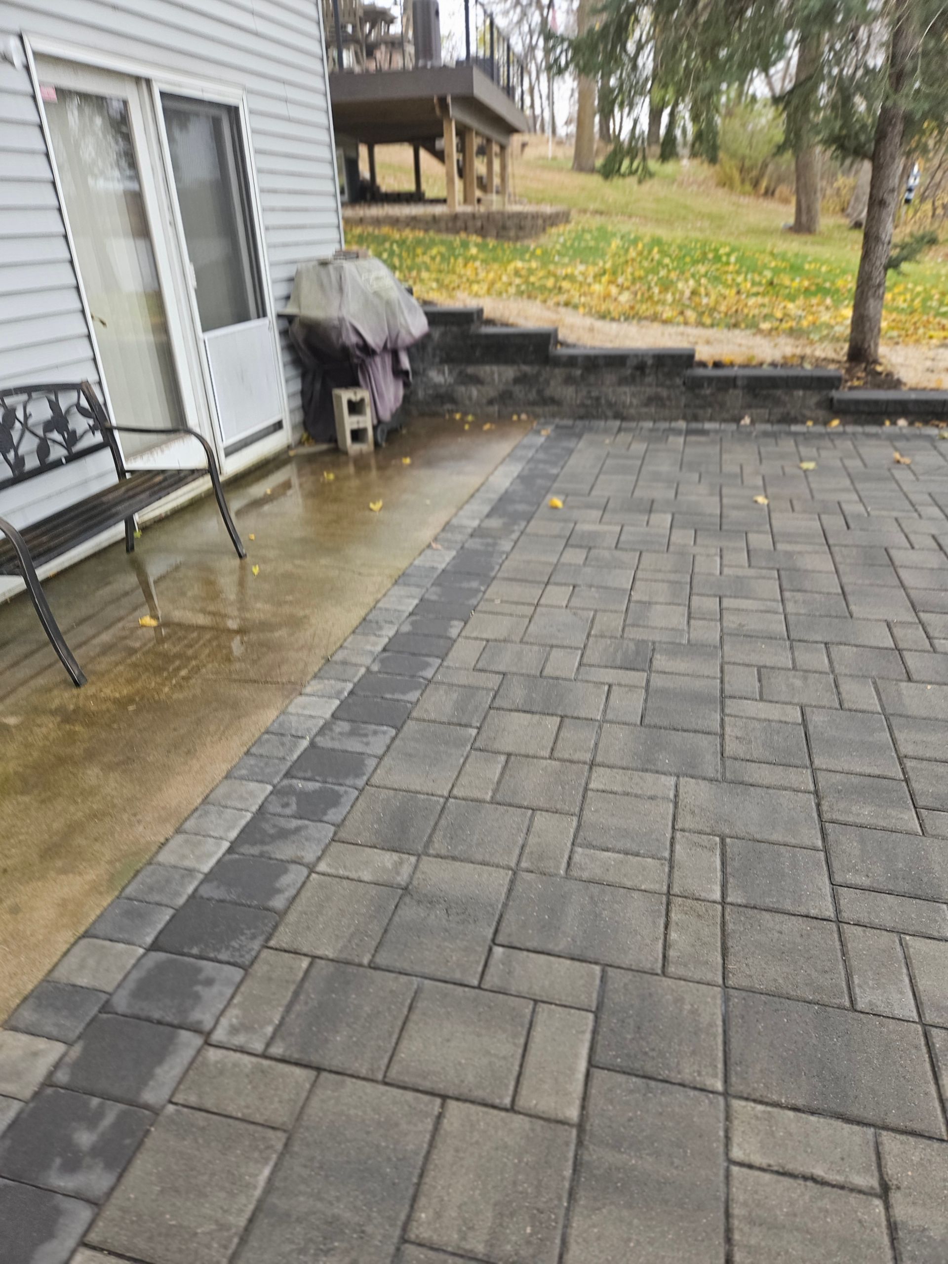 A stone patio with a gray paver border next to a house wall, a bench, and a tiered stone retaining wall in the backyard.