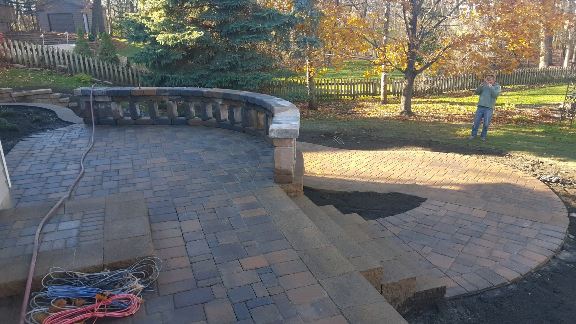 A stone patio under construction with a curved stone retaining wall and steps leading to a yard, featuring a standing person.