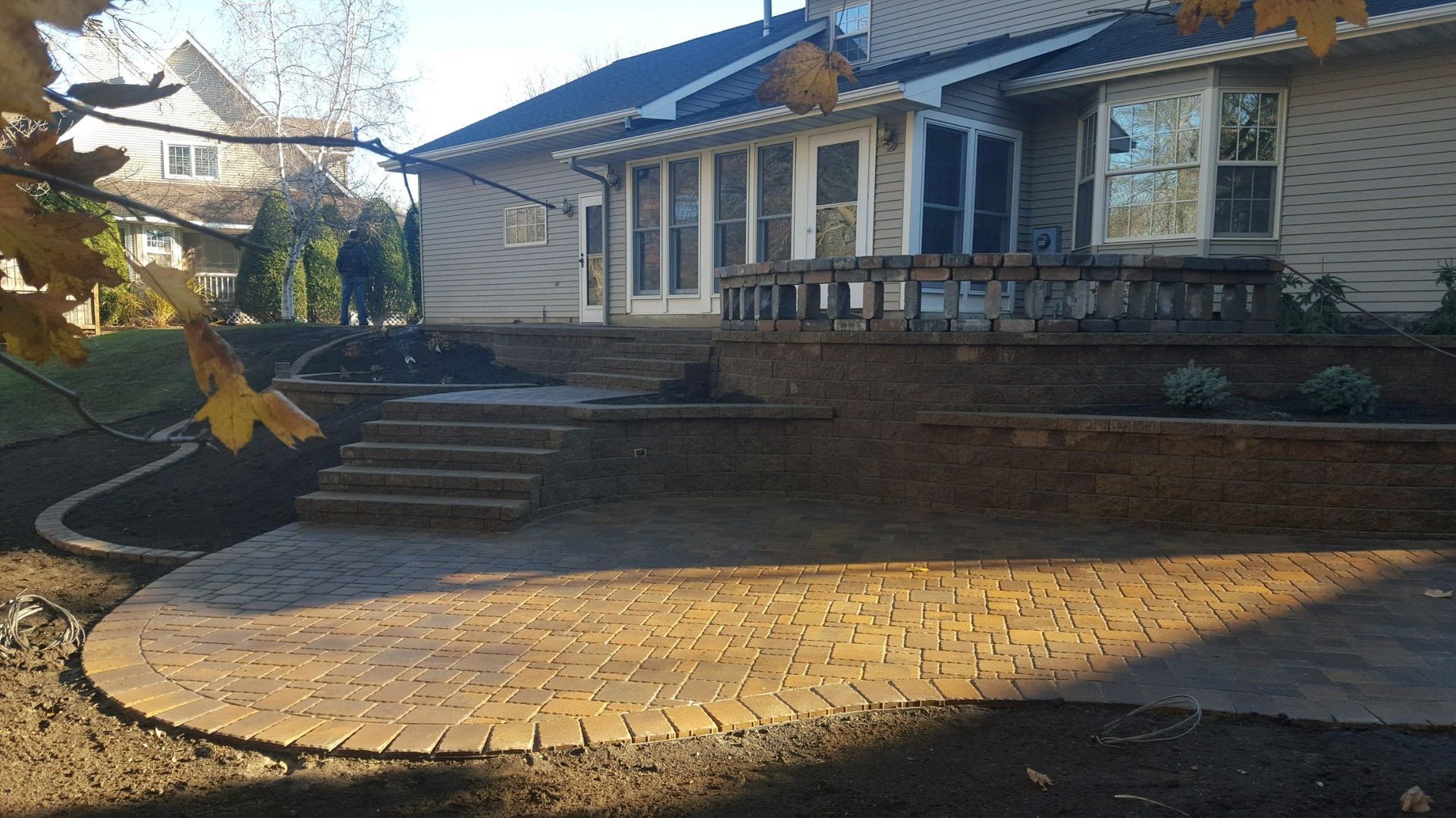 A wide shot of a backyard with a new brick paver patio, stone stairs, and a multi-level retaining wall against a house.