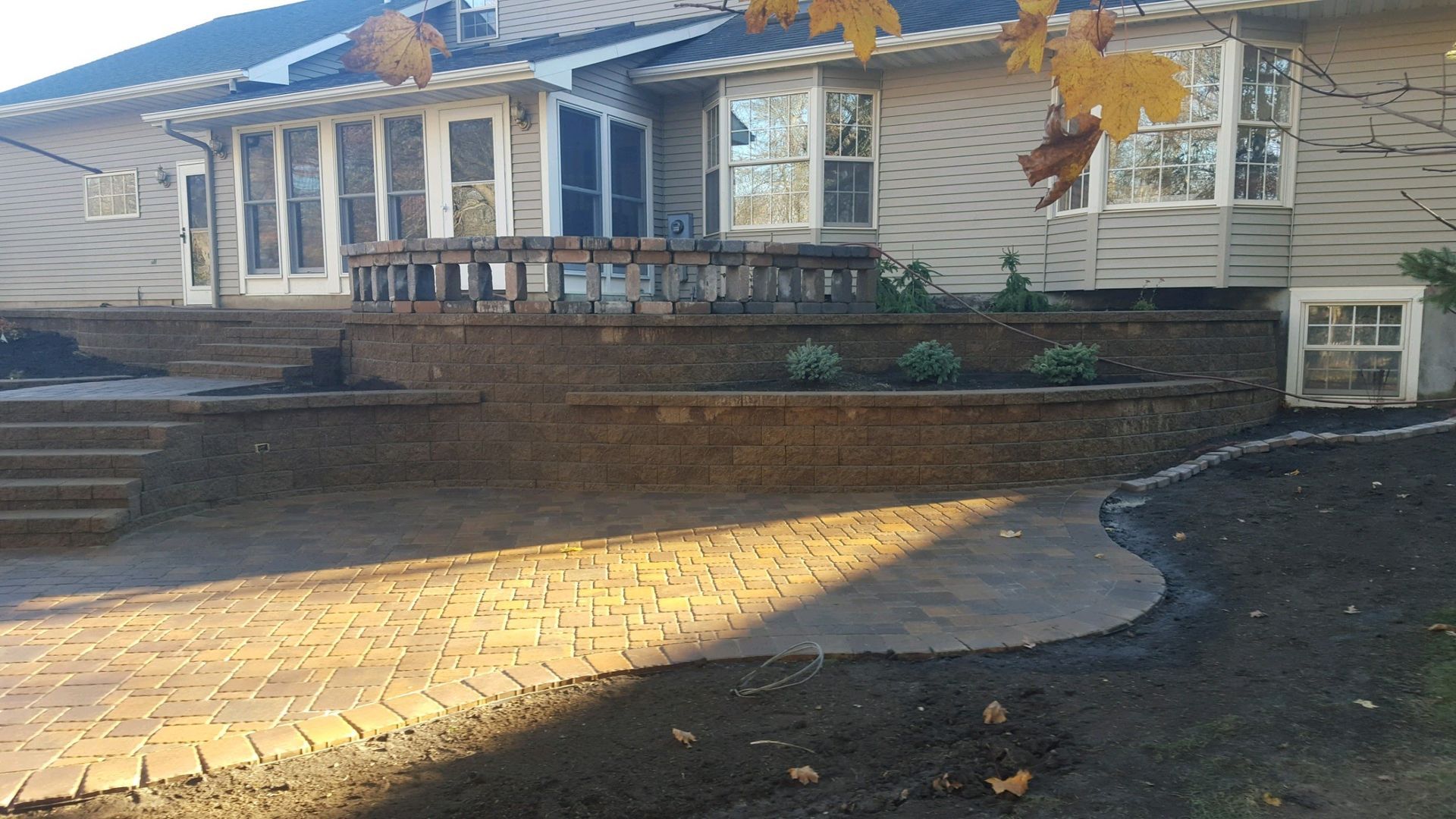 A house backyard featuring a tiered stone retaining wall, brick paver patio, and small plants in a sunny garden setting.