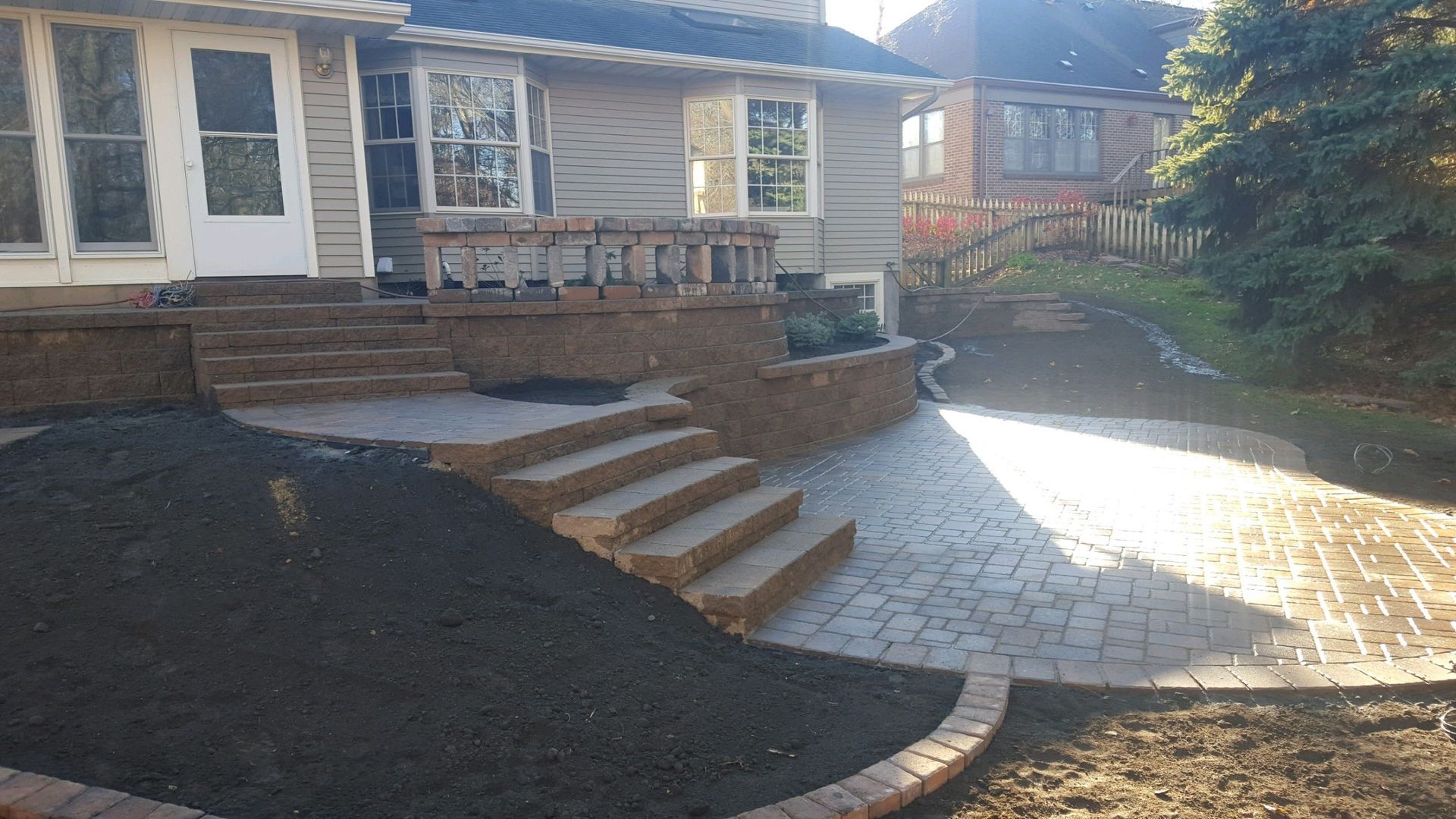 A beige house with a newly installed brown paver patio, stone retaining walls, and brick stairs leading to the lawn.