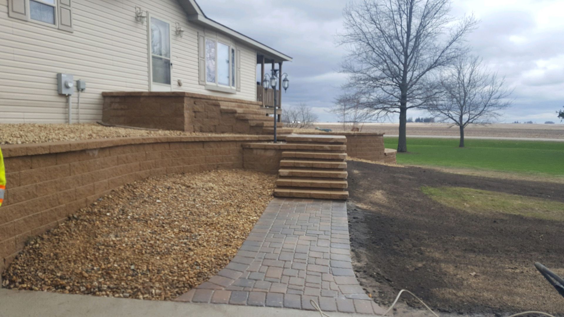 A paver walkway leads to a multi-tiered stone retaining wall and staircase in front of a tan siding house.
