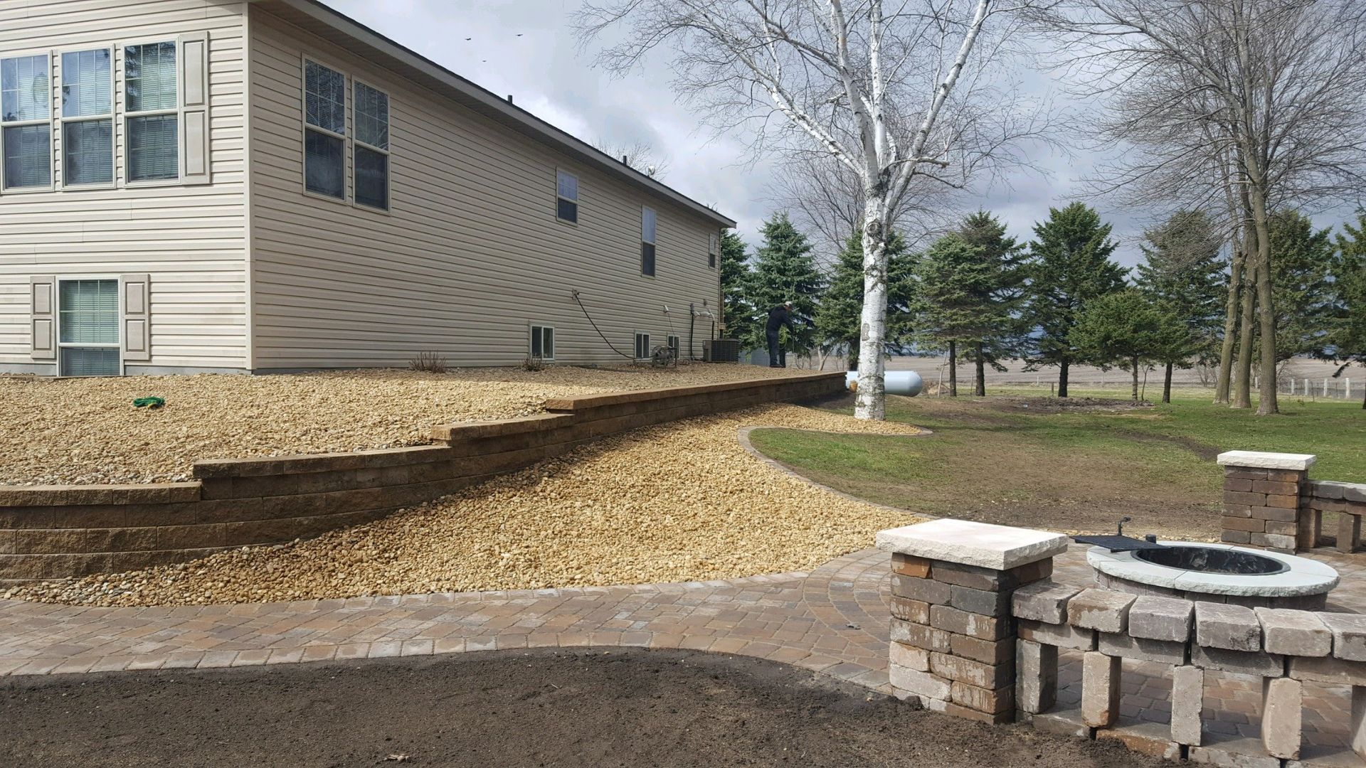 A backyard patio featuring a curved stone fire pit, a gravel-covered slope, and a tan house exterior under a cloudy sky.