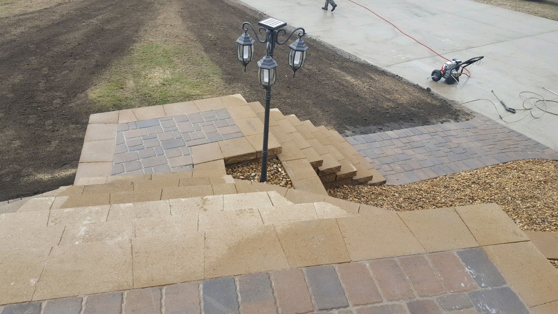 A multi-tiered brick patio featuring a central street-style lamppost surrounded by wood chips, set against a concrete drive.