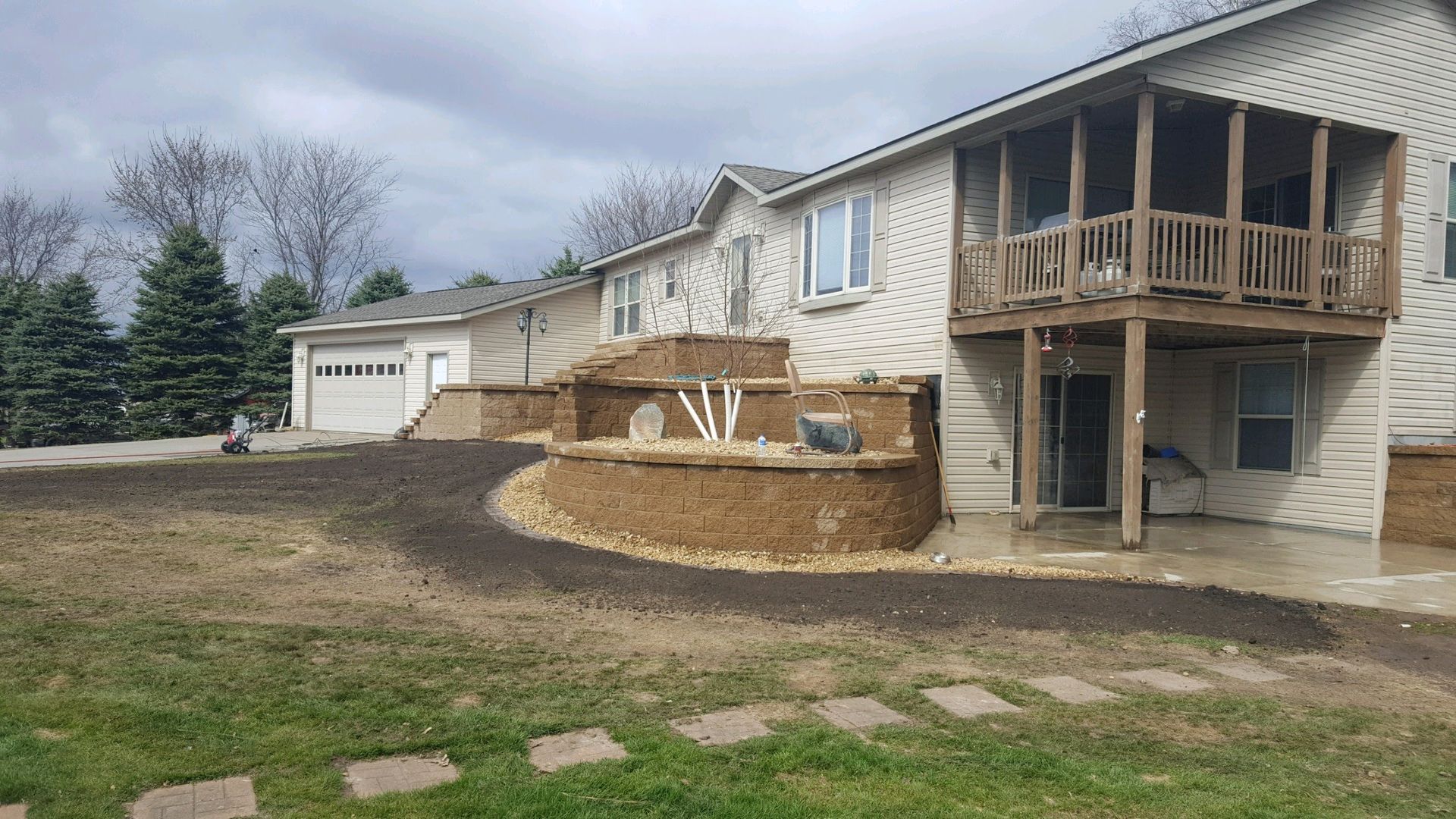A tiered retaining wall with fresh landscaping leads to a two-story house with a wooden deck and attached garage.