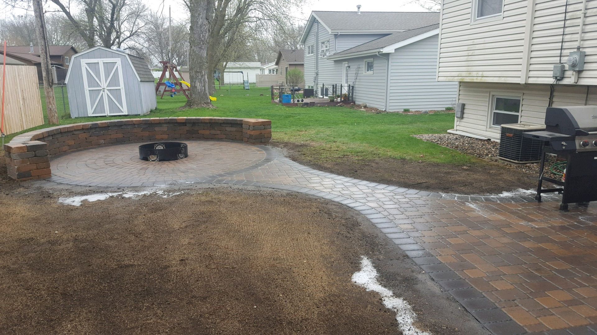 A newly installed stone patio with a curved seating wall, circular fire pit area, and a walkway leading to a house.