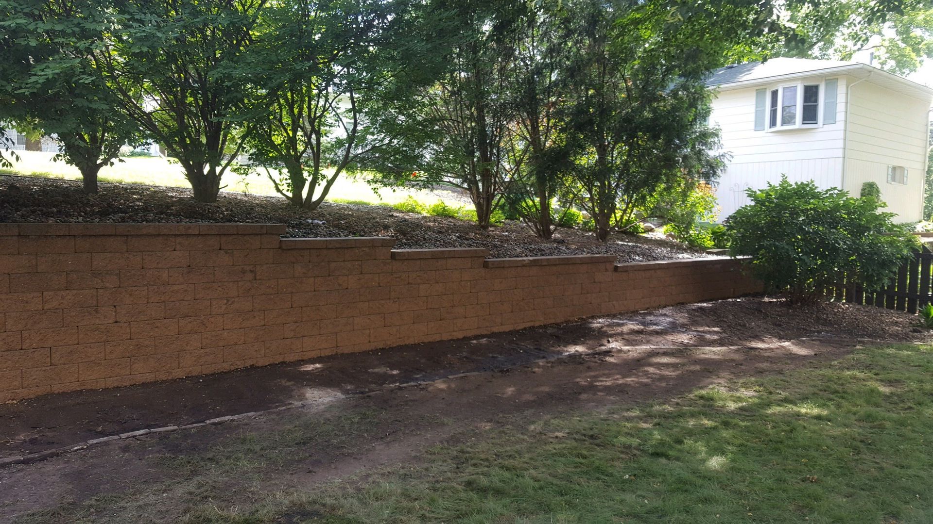 A tan stone retaining wall steps down a sloping grassy yard, with mature trees and a white house in the background.