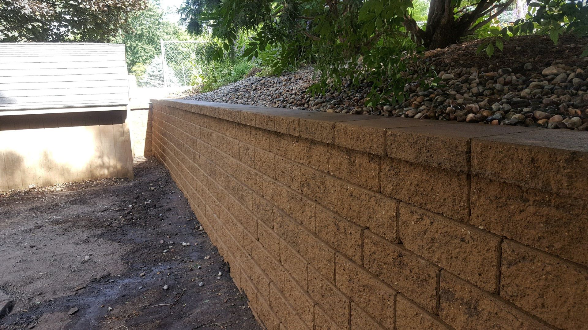 A tan, rectangular stone retaining wall borders a patch of dirt and a sloped, rock-covered garden bed.