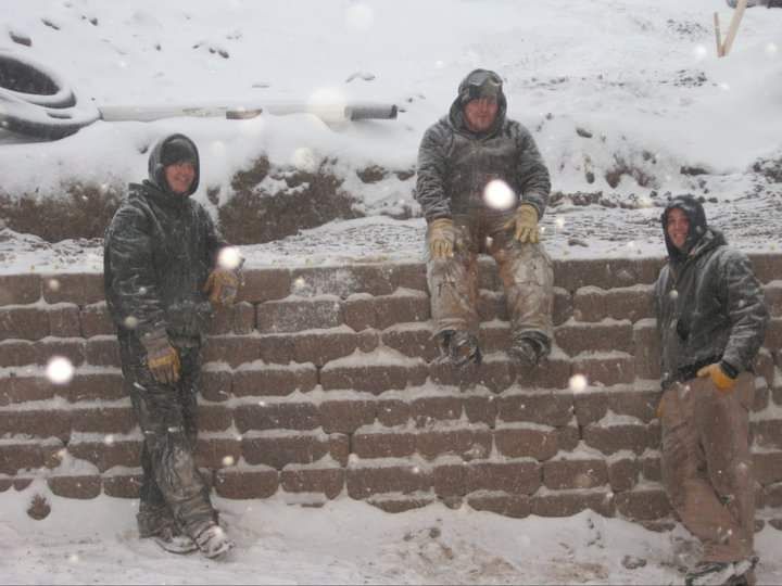 Three people in winter work gear standing or sitting along a stone retaining wall during a snowy day.