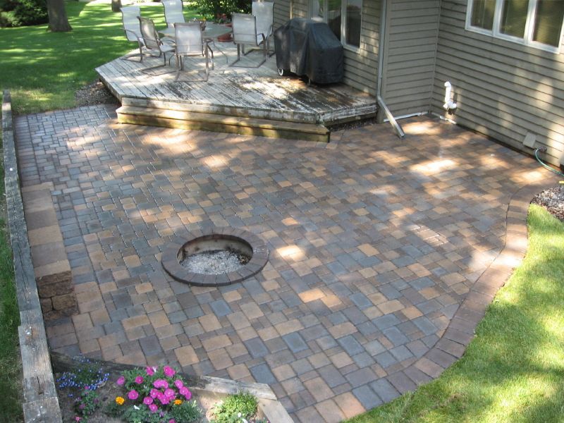 A paved patio with a circular fire pit, adjacent to a wooden deck and house, surrounded by lawn and landscaping.