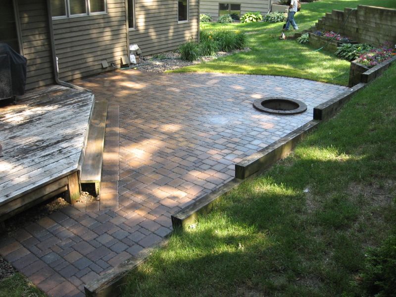 A multi-level backyard with a paved patio, a round stone fire pit, and a wooden deck attached to a house.