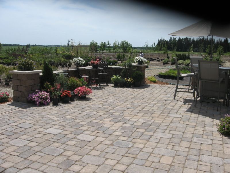 A paved patio with stone seating, potted flowers, and outdoor dining furniture, overlooking a large rural nursery.