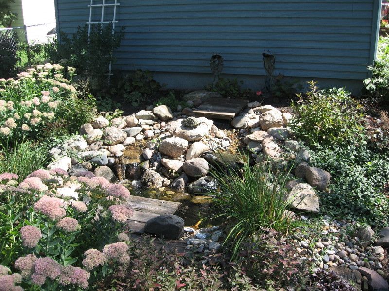 A backyard water feature with a stone-lined stream and pond, surrounded by lush green foliage and flowering plants.